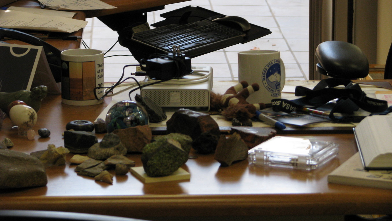 Prof. Brown's desk with several asteroid and other samples. The SciSolSys pallasite is in the plastic case in the right foreground; the SciSolSys coffee mug in the left background.